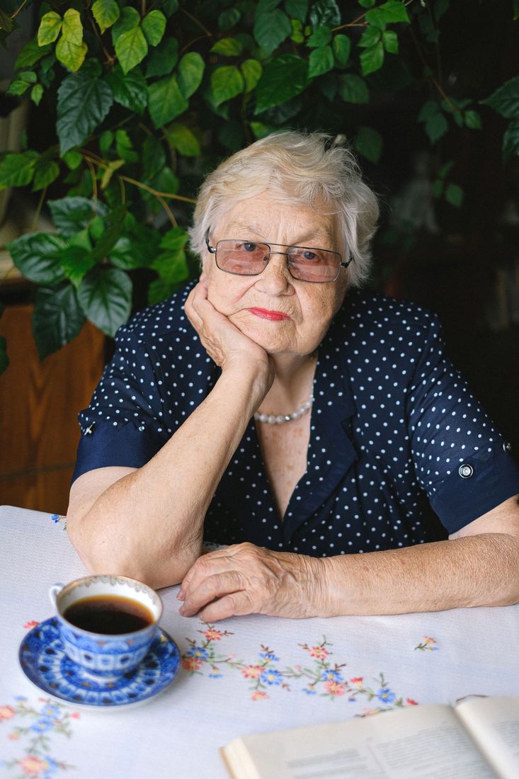 An older woman sits at her table with a cup of tea, chin resting on her hand